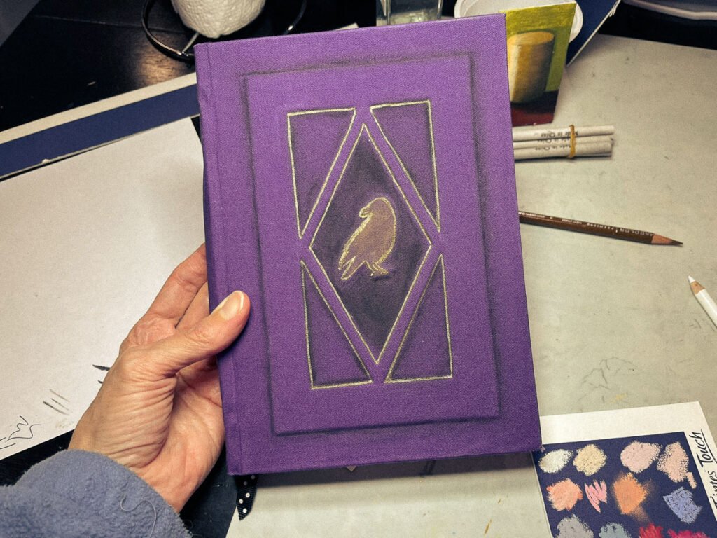 Hand holding a handmade sketchbook with a purple cover and a gold-lined raven design on the front.