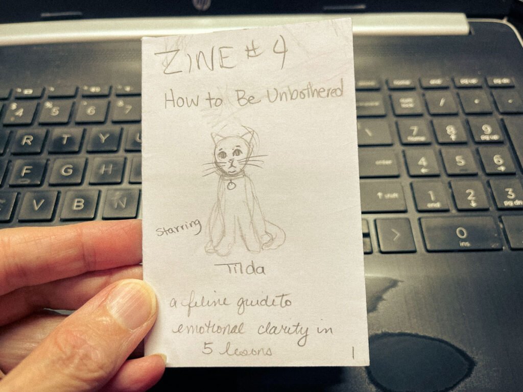 Hand holding a folded paper mockup of a zine titled “Zine #4: How to Be Unbothered,” featuring a pencil sketch of a cat named Tilda.