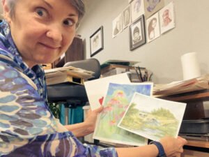 Artist smiling while holding up a watercolor flower painting and a landscape sketch in a cozy, art-filled studio