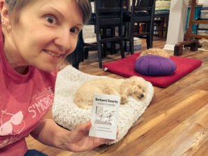 Anni holds up her mini zine titled “Barkyard Security: Unleashed & Unqualified” while smiling. Two small, curly-haired dogs rest in the background.