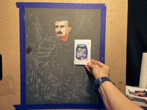 A hand holding a small illustrated zine titled “How to Catch a Story” in front of a larger unfinished oil pastel portrait of a male paladin in armor.
