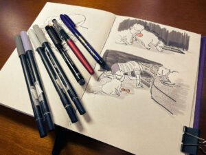 Open sketchbook on a wooden desk showing grayscale dog cartoons; four Tombow gray brush markers and three black brush pens rest across the left page.