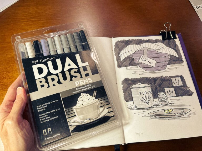 Hand holding a Tombow Dual Brush grayscale marker set beside a sketchbook page shaded with the same cool-gray tones.