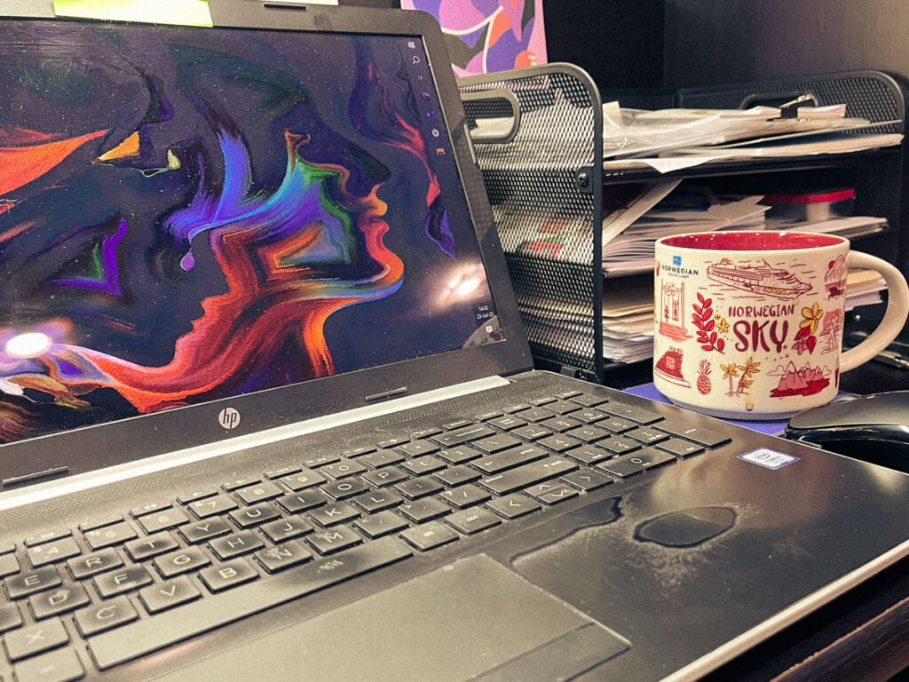 Laptop on a desk with a colorful abstract face design on the screen, next to a Norwegian Sky cruise souvenir coffee mug and a black wire organizer filled with papers.