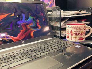 Laptop on a desk with a colorful abstract face design on the screen, next to a Norwegian Sky cruise souvenir coffee mug and a black wire organizer filled with papers.