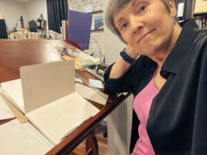 Artist at a cluttered drafting table, sketchbook open with simple line drawings, smiling slightly at the camera in a casual creative workspace.