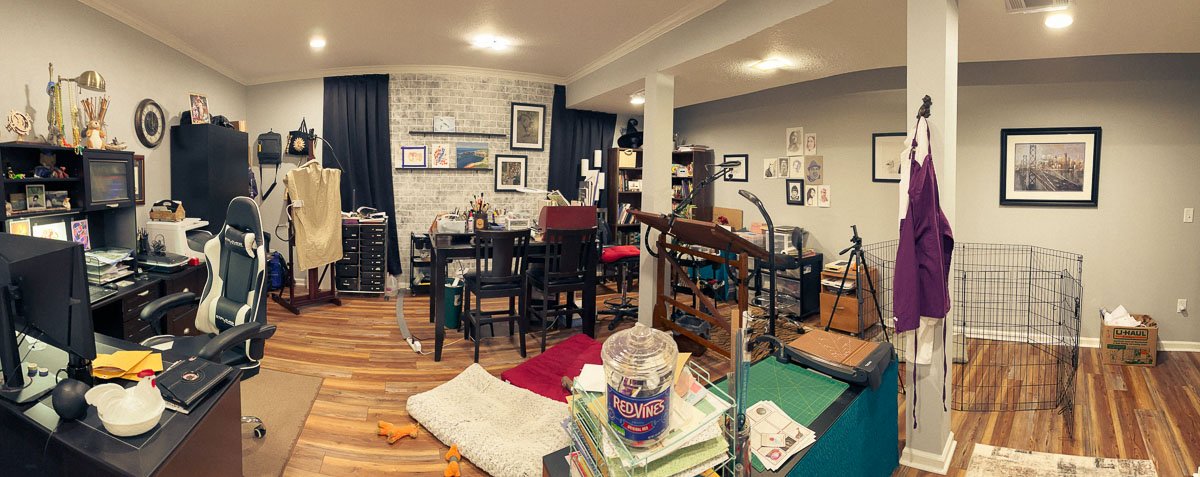 Panoramic view of a lived-in home art studio with drafting tables, computer workstation, art supplies, and creative clutter across the space