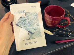 Hand holding a folded zine titled “The Shopping List” with illustrated cover, beside a red Chico State mug, reading glasses, a pen, and a beaded necklace on a desk.