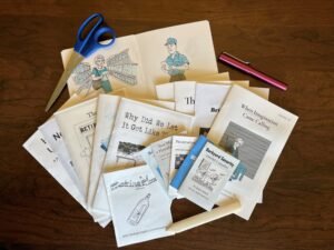 A spread of handmade zines fanned out on a wooden table, with scissors, a pink pen, and a bone folder beside them.