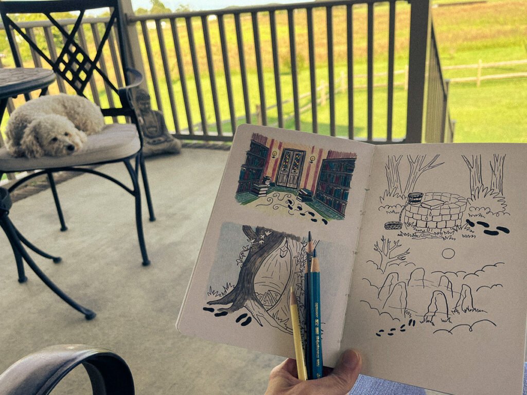 Hand holding a sketchbook of zine illustrations with pencils on a patio table, while a small white dog rests on a chair nearby.