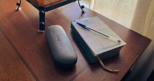 A small notepad and pen rest on a bedside table beside a glasses case and lamp, waiting to capture ideas that come before sleep or upon waking.