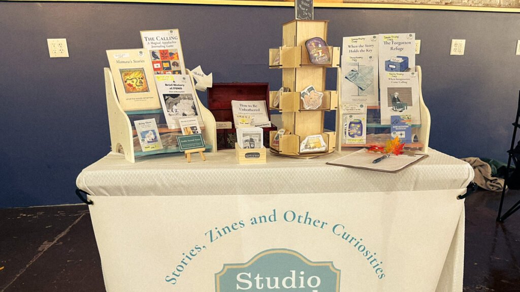 Studio Second Street zine display at Johnson City Zine Fest, set against white brick wall.
