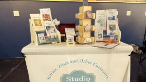 Studio Second Street zine display at Johnson City Zine Fest, set against white brick wall.