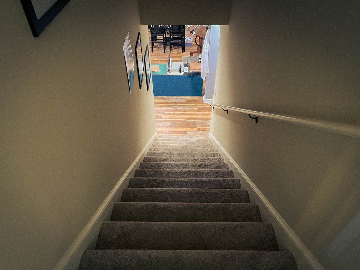 View from the top of a carpeted staircase leading down to a warmly lit basement studio — a quiet moment of returning to creative life after weeks away.