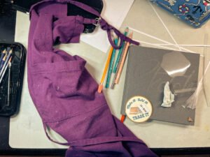 A purple paint-splattered apron rests on a messy worktable beside pencils, a sketchbook, and paper scraps — the quiet evidence of my return to the studio after a long pause.