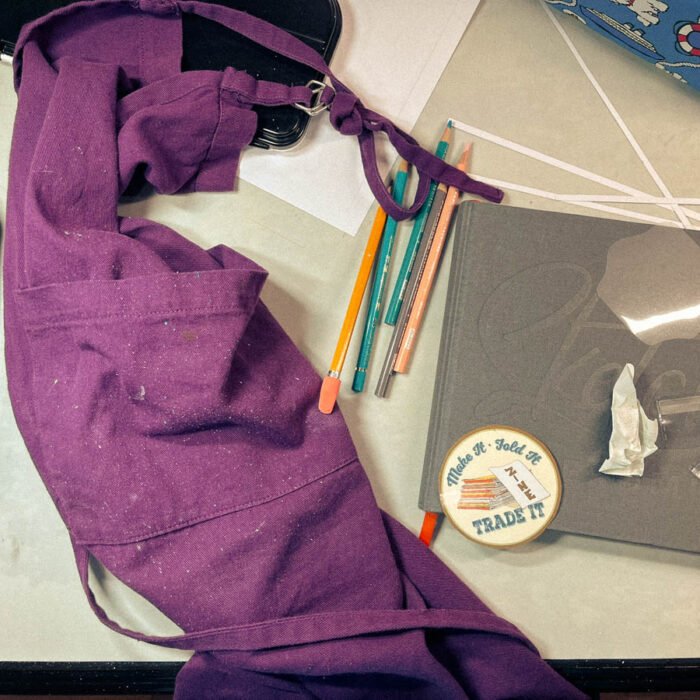 A purple paint-splattered apron rests on a messy worktable beside pencils, a sketchbook, and paper scraps — the quiet evidence of my return to the studio after a long pause.