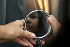 Hands holding a small round mirror with a blurred face reflected inside, against a soft neutral background with warm light.
