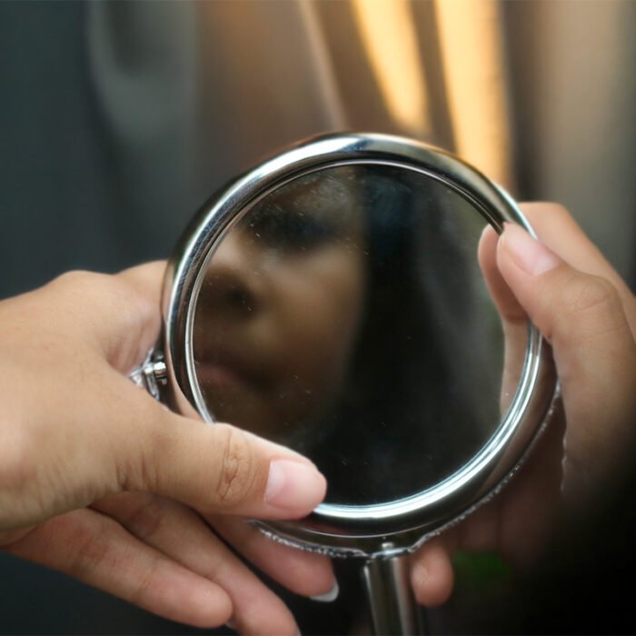 Hands holding a small round mirror with a blurred face reflected inside, against a soft neutral background with warm light.