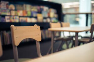 A soft-focus photo of a library reading area, with an empty wooden chair in the foreground and shelves of colorful picture books blurred in the background.