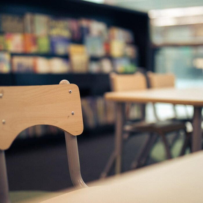 A soft-focus photo of a library reading area, with an empty wooden chair in the foreground and shelves of colorful picture books blurred in the background.