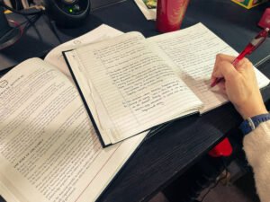 Open book and notebook on a desk, with handwritten notes and a hand writing with a red pen, documenting research and reflections for an altered book project.