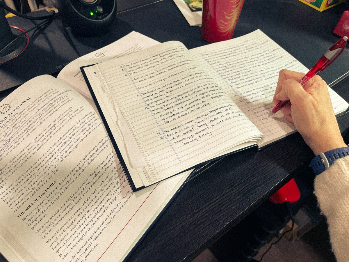Open book and notebook on a desk, with handwritten notes and a hand writing with a red pen, documenting research and reflections for an altered book project.