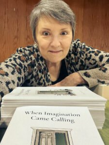 A woman leaning forward behind a stack of books titled When Imagination Came Calling, with the book cover visible in the foreground.