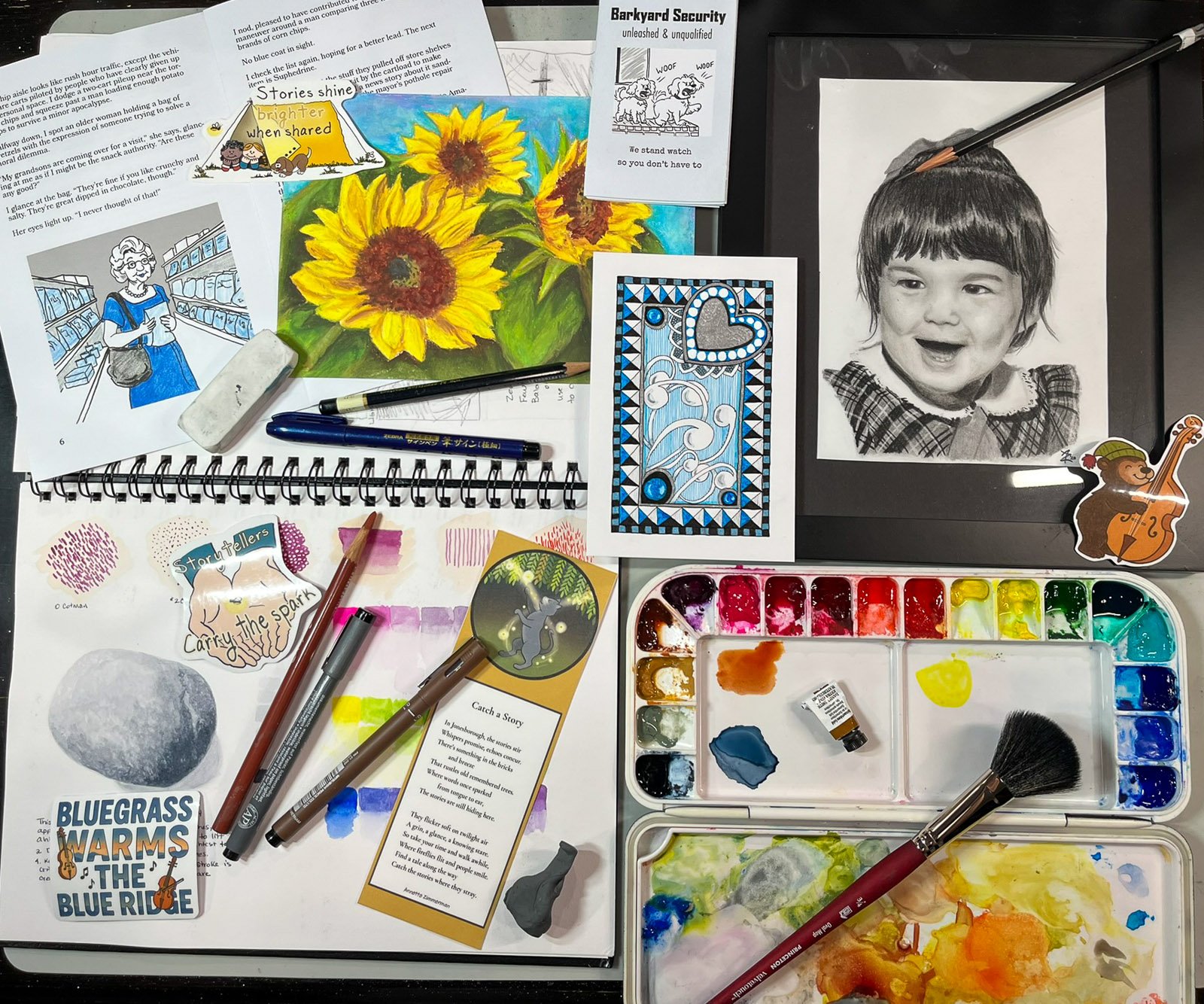 A creative worktable filled with sketchbooks, watercolor paints, drawings, zines, printed story pages, pens, brushes, and small art pieces.