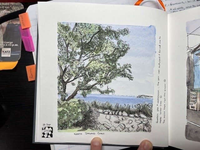 Sketchbook page showing a pen and watercolor study of the North Ireland coast, with a tree, stone wall, grasses, sea, and pale blue sky.