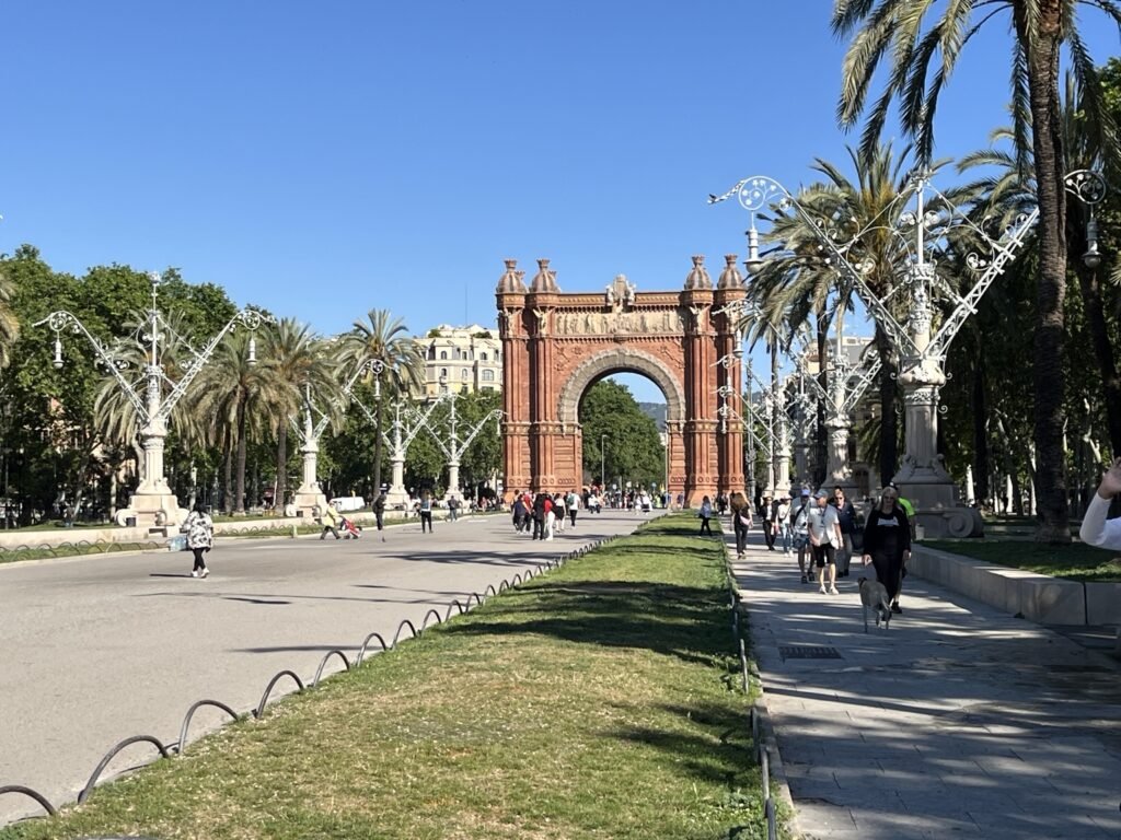 Reference photo of Barcelona’s Arc de Triomf seen from a wide pedestrian avenue lined with palm trees and ornate street lamps.