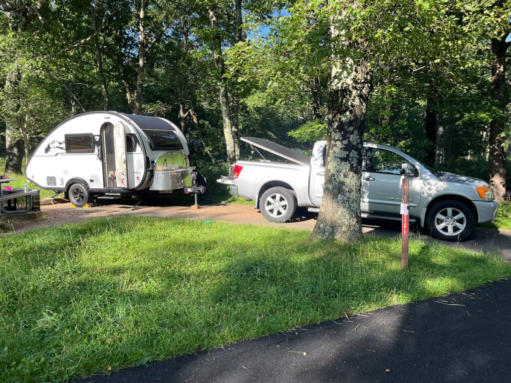 A white Tab 400 trailer and silver pickup truck parked at a shaded campsite surrounded by green trees.