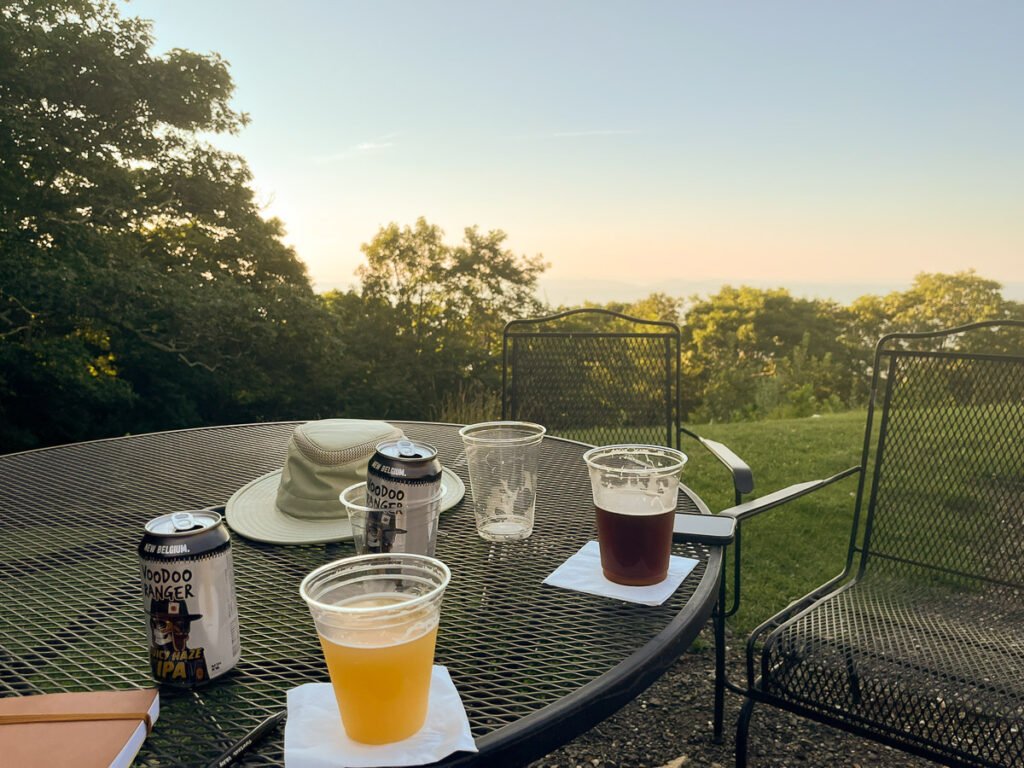 Plastic cups of beer, two cans, and a sun hat sit on a metal patio table overlooking trees and a hazy mountain ridge.
