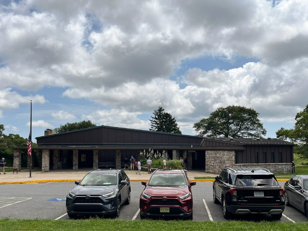 Stone-and-dark-wood Byrd Visitor Center beneath large summer clouds, with several cars parked in front.