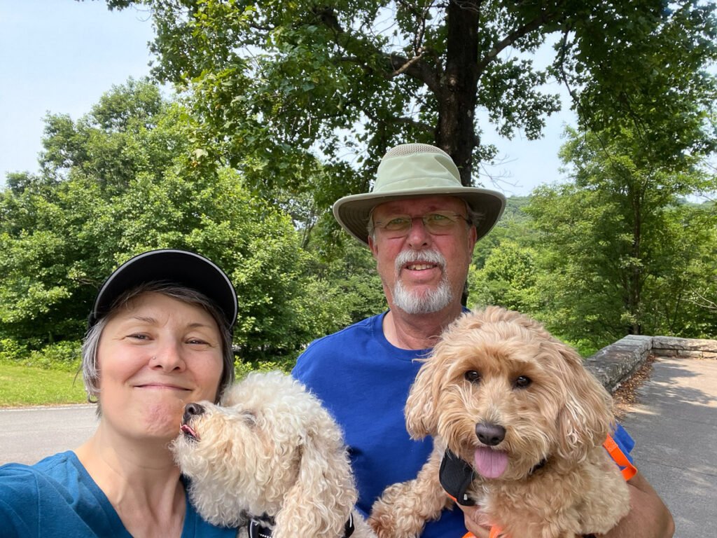 A smiling couple poses outdoors with two curly-haired dogs against a backdrop of green trees and mountains.