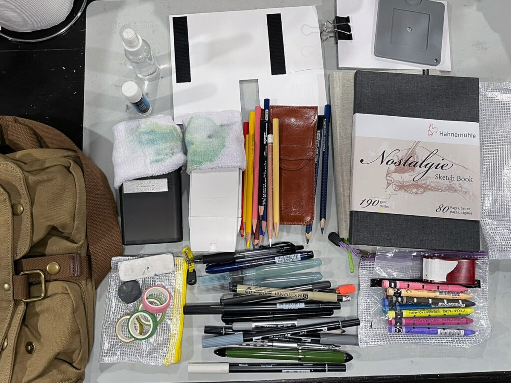A large assortment of travel art supplies arranged beside a brown shoulder bag, including pens, pencils, watercolor palettes, brushes, colored pencils, crayons, tape, and a gray sketchbook.