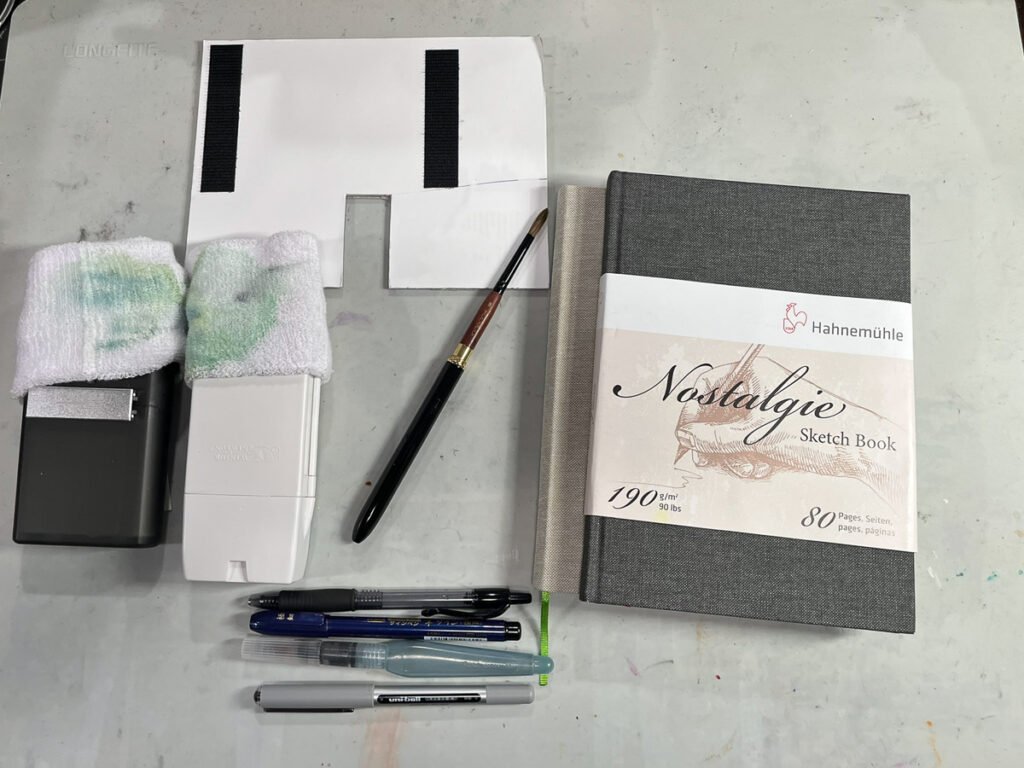 A reduced travel art kit containing two watercolor palettes, one brush, a gray sketchbook, several pens, and two waterbrushes.