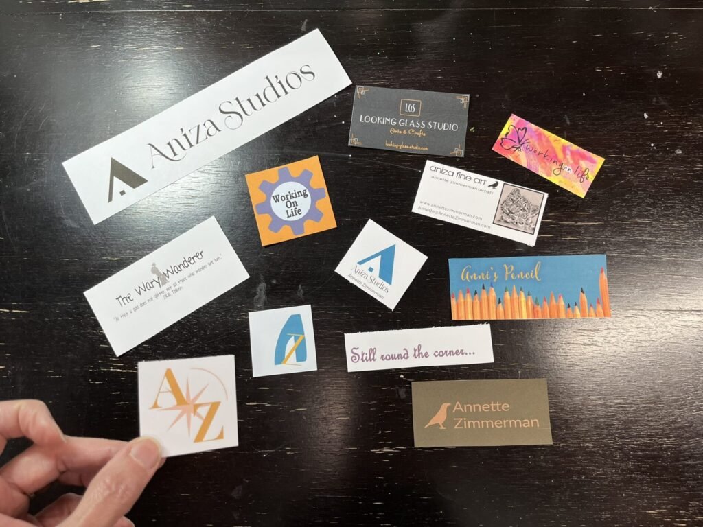 A hand holds an old Aniza logo card above a dark wooden table scattered with business cards and logos from ten years of creative projects and blogs.