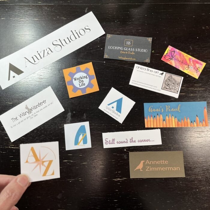 A hand holds an old Aniza logo card above a dark wooden table scattered with business cards and logos from ten years of creative projects and blogs.