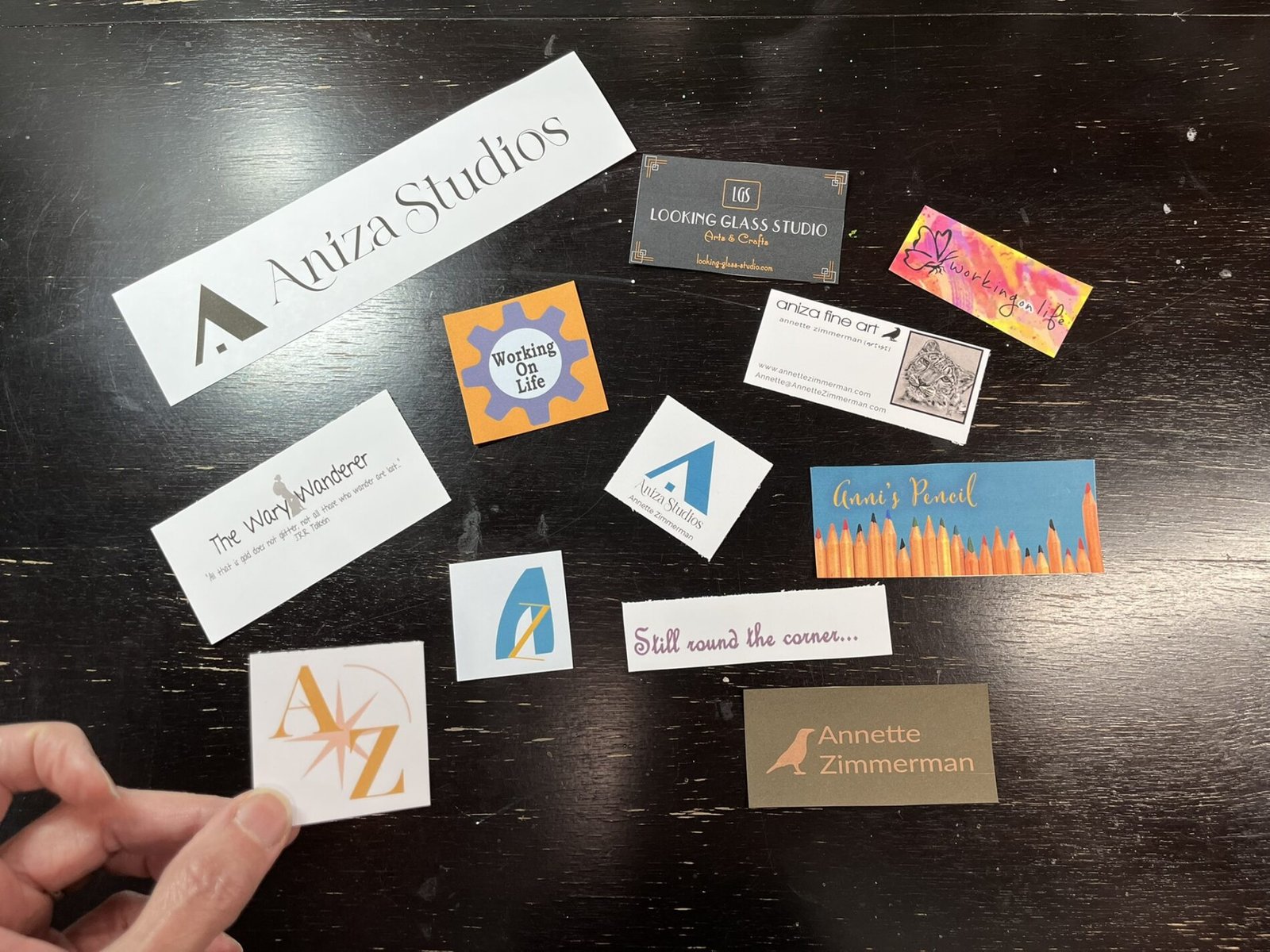 A hand holds an old Aniza logo card above a dark wooden table scattered with business cards and logos from ten years of creative projects and blogs.