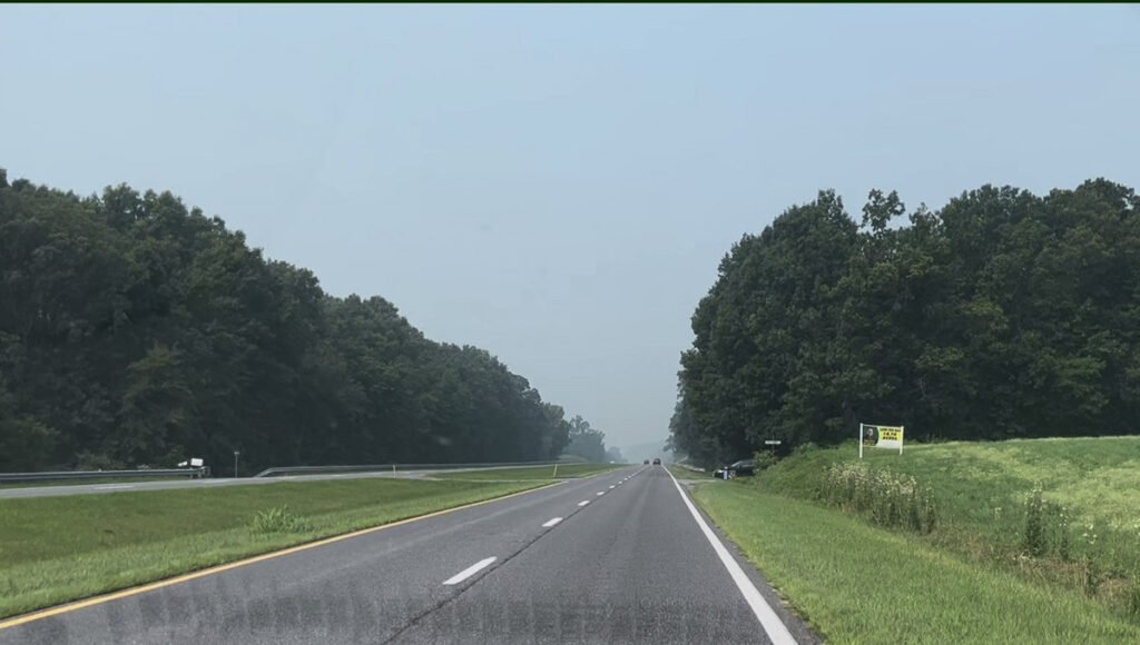 Hazy divided highway bordered by green trees and fields, with wildfire smoke reducing visibility in the distance.