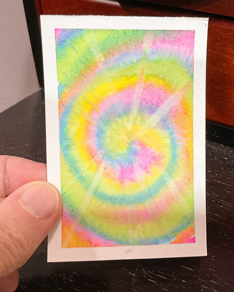 A hand holds a small card painted with concentric rings of pink, yellow, green, blue, and lavender watercolor.