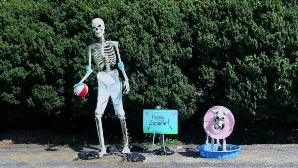 Large decorative skeleton in swim trunks holding a beach ball beside a smaller skeleton dog standing in a blue kiddie pool.