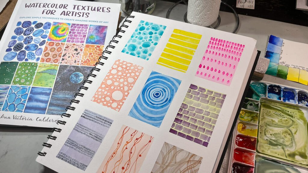 A sketchbook page filled with nine rectangular watercolor texture exercises beside the book Watercolor Textures for Artists and a watercolor palette.