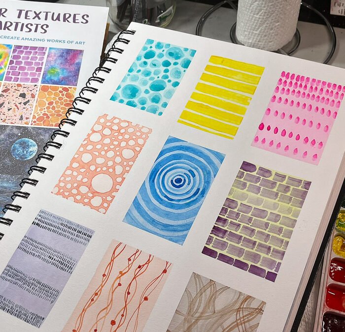A sketchbook page filled with nine rectangular watercolor texture exercises beside the book Watercolor Textures for Artists and a watercolor palette.