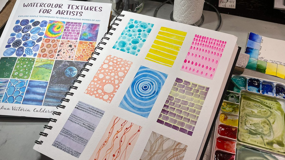 A sketchbook page filled with nine rectangular watercolor texture exercises beside the book Watercolor Textures for Artists and a watercolor palette.