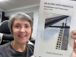 Anni Zimmerman smiles while holding the August 2026 Realmscapes Dispatch newsletter, titled “Foundations and Frameworks,” featuring an illustration of a person standing on a steel beam above a skyscraper.