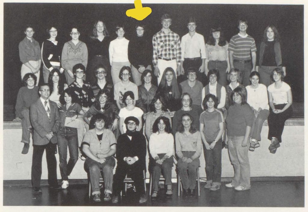 Black-and-white group photograph of a high school drama club posed on a stage, with a yellow arrow pointing to Anni in the back row.