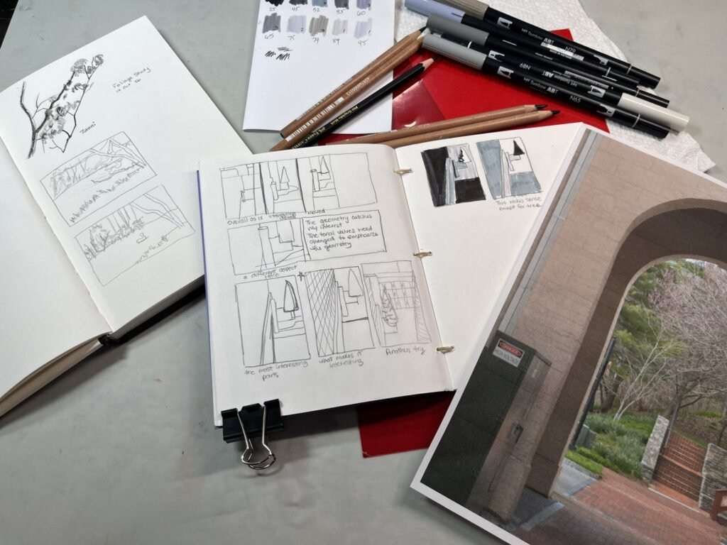 Open sketchbooks with architectural thumbnails, handwritten notes, and tonal studies beside pencils, gray markers, swatches, and a reference photograph of a brick archway.