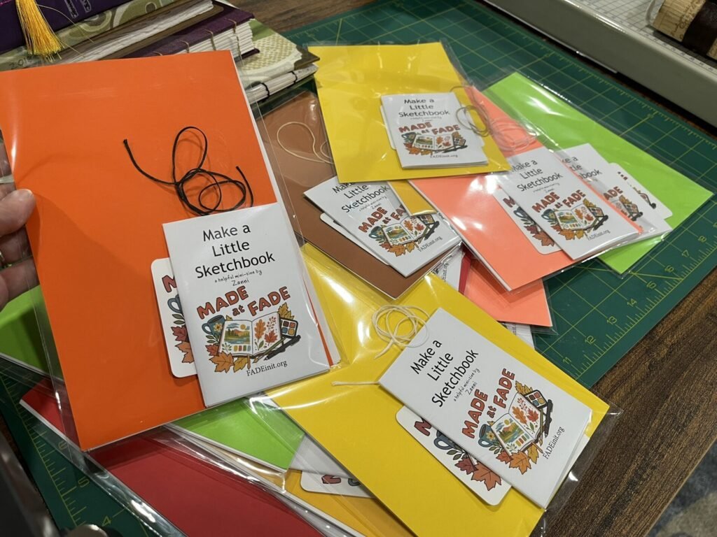 Sketchbook-making kits in clear bags on a cutting mat, with colorful covers, binding thread, “Made at FADE” stickers, and “Make a Little Sketchbook” instruction zines.